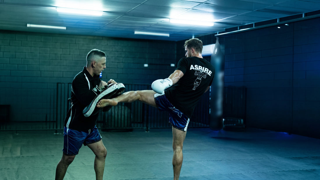 ASPIRE. athlete throwing a barefoot Muay Thai kick into pads held by his coach, ASPIRE back print visible